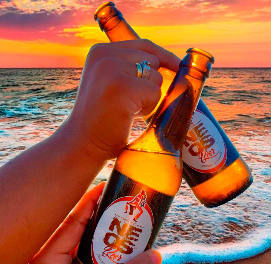 Manos levantando botellas de cerveza NEOS al atardecer en la playa, con el mar y el cielo colorido de fondo.