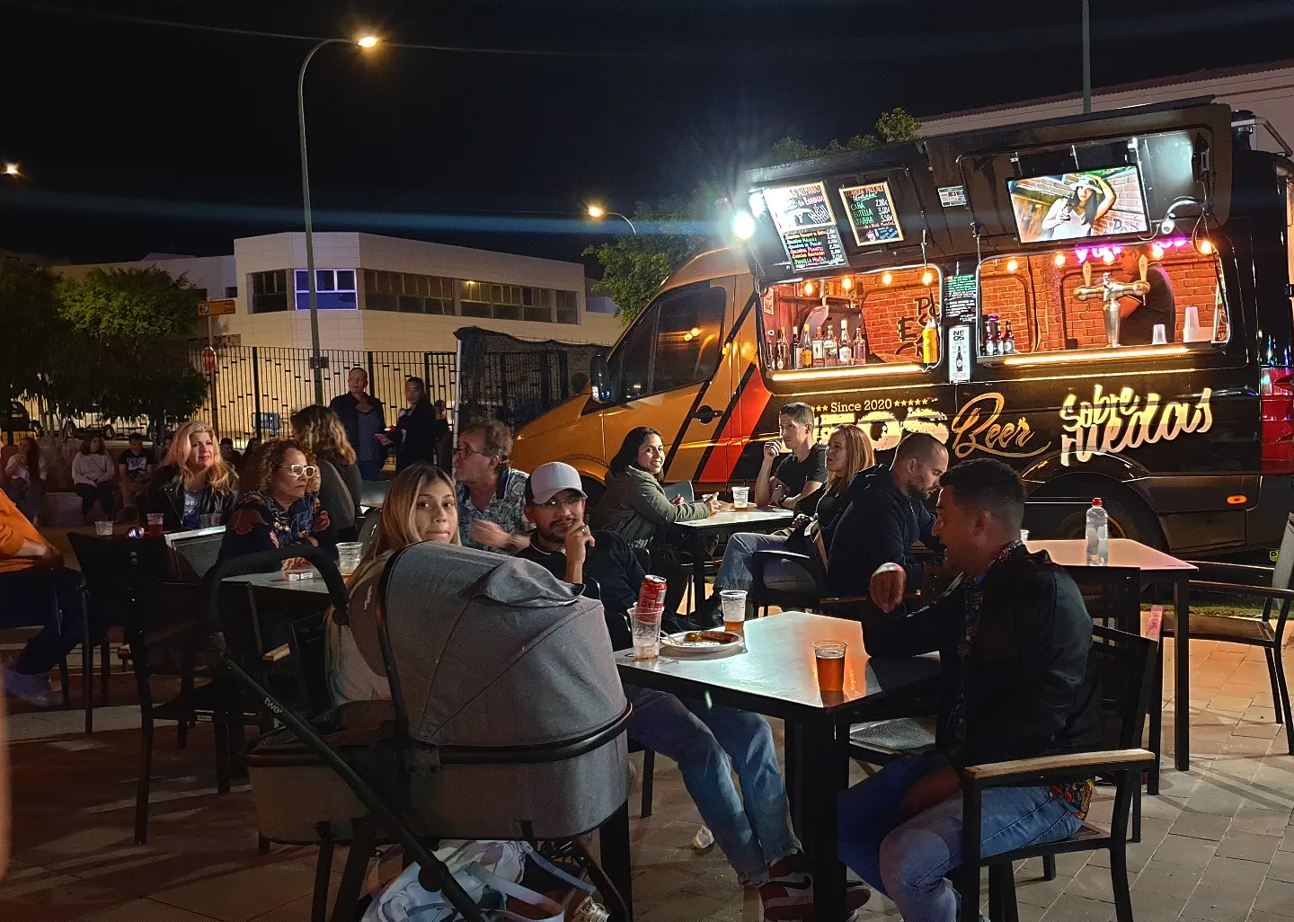 Clientes disfrutando de una noche en un food truck de cervezas, con mesas al aire libre y un ambiente animado. Se observan bebidas y un menú iluminado en el camión.